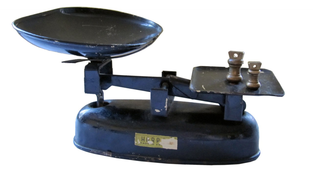 A vintage, dark blue balance scale with a large, shallow pan on the left side and a smaller, flat platform on the right. The platform currently holds two brass weights of different sizes. The base of the scale is rounded, with a worn label that reads "HARP MADE IN ENGLAND." The scale shows signs of wear, with scuffs and chipped paint, indicating it has been well-used. The design is simple and functional, representative of traditional mechanical weighing scales used in the past.