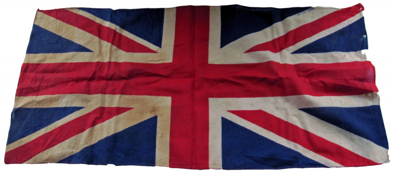 A worn Union Jack flag, displaying the red, white, and blue colours of the United Kingdom. The fabric shows signs of age, with visible creases, stains, and a few small tears along the edges. The colours appear slightly faded, indicating that the flag has likely been exposed to the elements or used extensively over time.