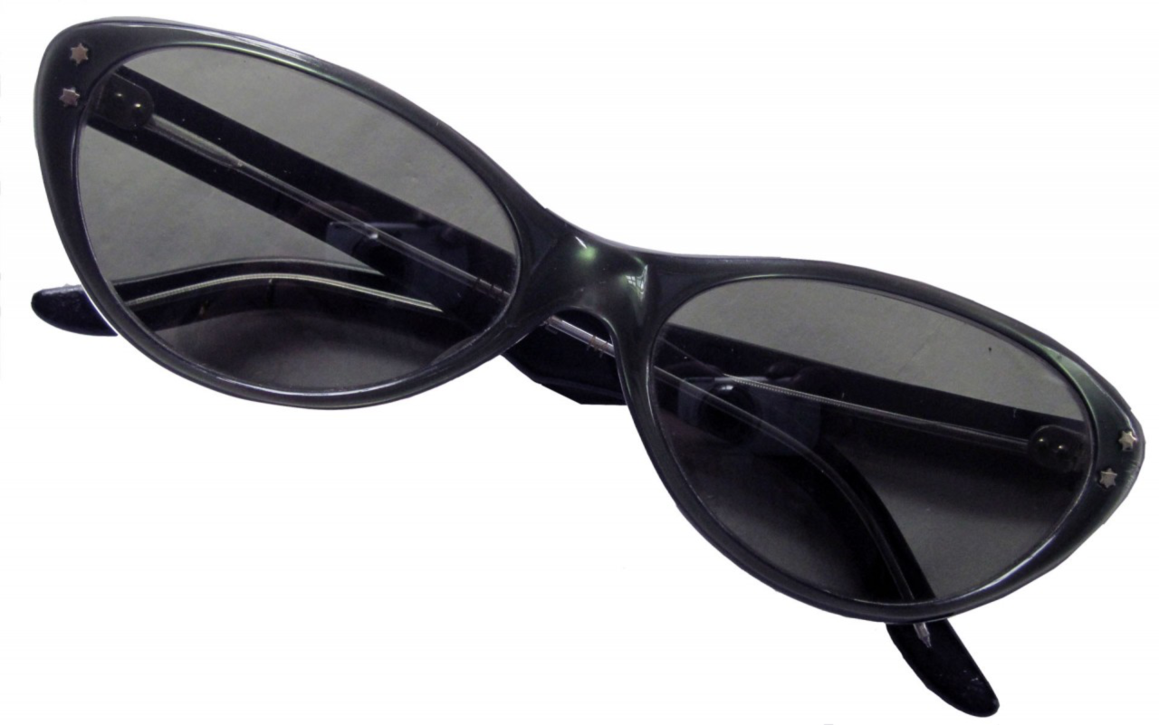 A pair of vintage cat-eye sunglasses with dark, slightly tinted lenses and black plastic frames. The frames are stylishly pointed at the corners, typical of the cat-eye design popular in the mid-20th century. Small decorative star-shaped studs adorn the outer edges of the frames, adding a touch of flair. The lenses are in good condition, though the frames show minor signs of wear, suggesting they have been used.