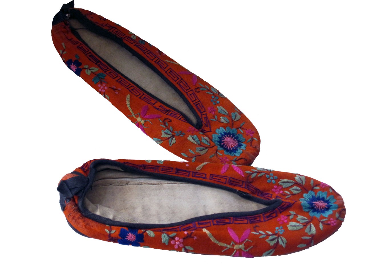 A pair of traditional embroidered slip-on shoes, featuring a vibrant orange fabric adorned with intricate floral patterns in shades of blue, pink, green, and purple. The embroidery is detailed, with flowers and leaves scattered across the surface, and a decorative geometric border in black and orange running along the edges of the shoe openings. The shoes have a soft, flat sole and a slightly rounded toe. The inner lining appears worn, suggesting they have been used, but the exterior remains colourful and well-preserved.